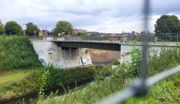 Blick auf Br&uuml;cke mit begr&uuml;nten Ufern am Fluss und Zaun im Vordergrund