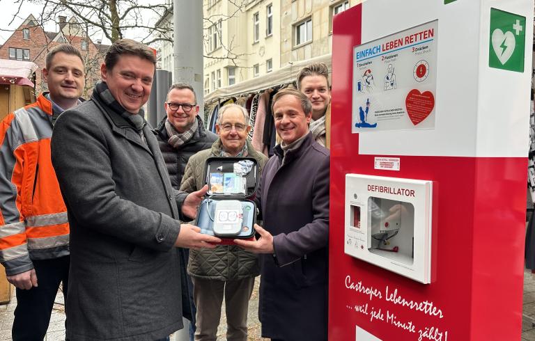 Sechs Personen, die vor einer AED-S&auml;ule stehen und einen Defibrillator pr&auml;sentieren